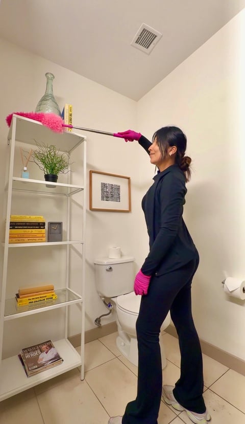 Woman in black outfit with pink gloves using a feather duster to clean bathroom shelves while standing next to a toilet.